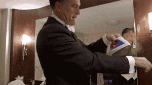Animated GIF: a man in a dark suit, in front of a mirror in a room, irons the sleeve of the shirt he is wearing, helped by another person. No text in the image.